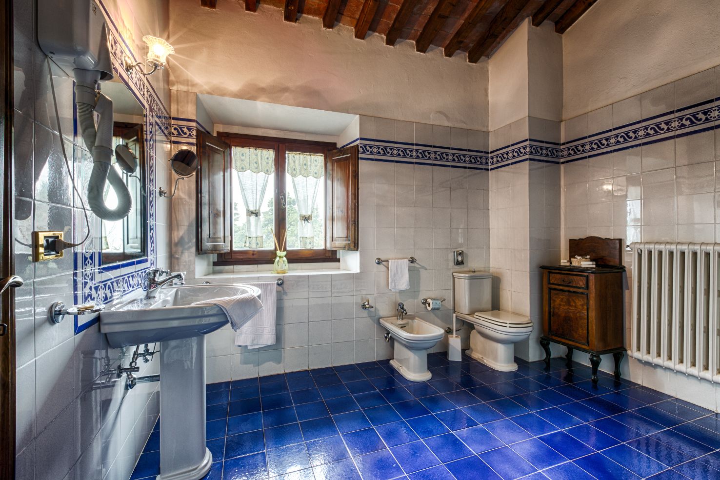 A bathroom with a sink, two toilets, a window with curtains, and a radiator.