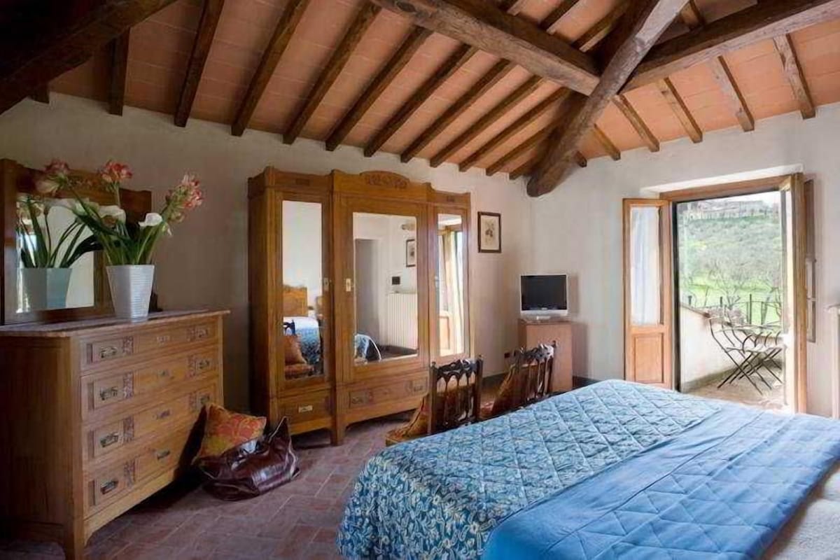 Ein Schlafzimmer mit Bett, einem Holzschrank und einem Fernseher. Es gibt einen Balkon mit Stuhl und Tisch.