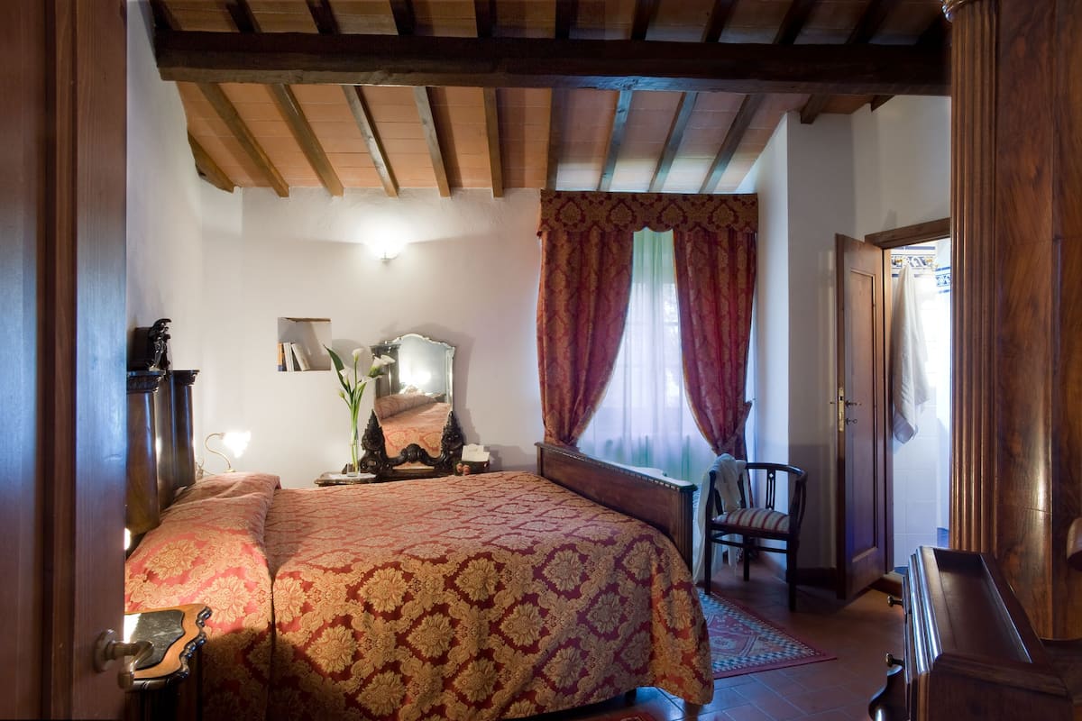 Un dormitorio tradicional con techo de madera, una cama con colcha estampada, una silla y una mesita de noche con lámpara.