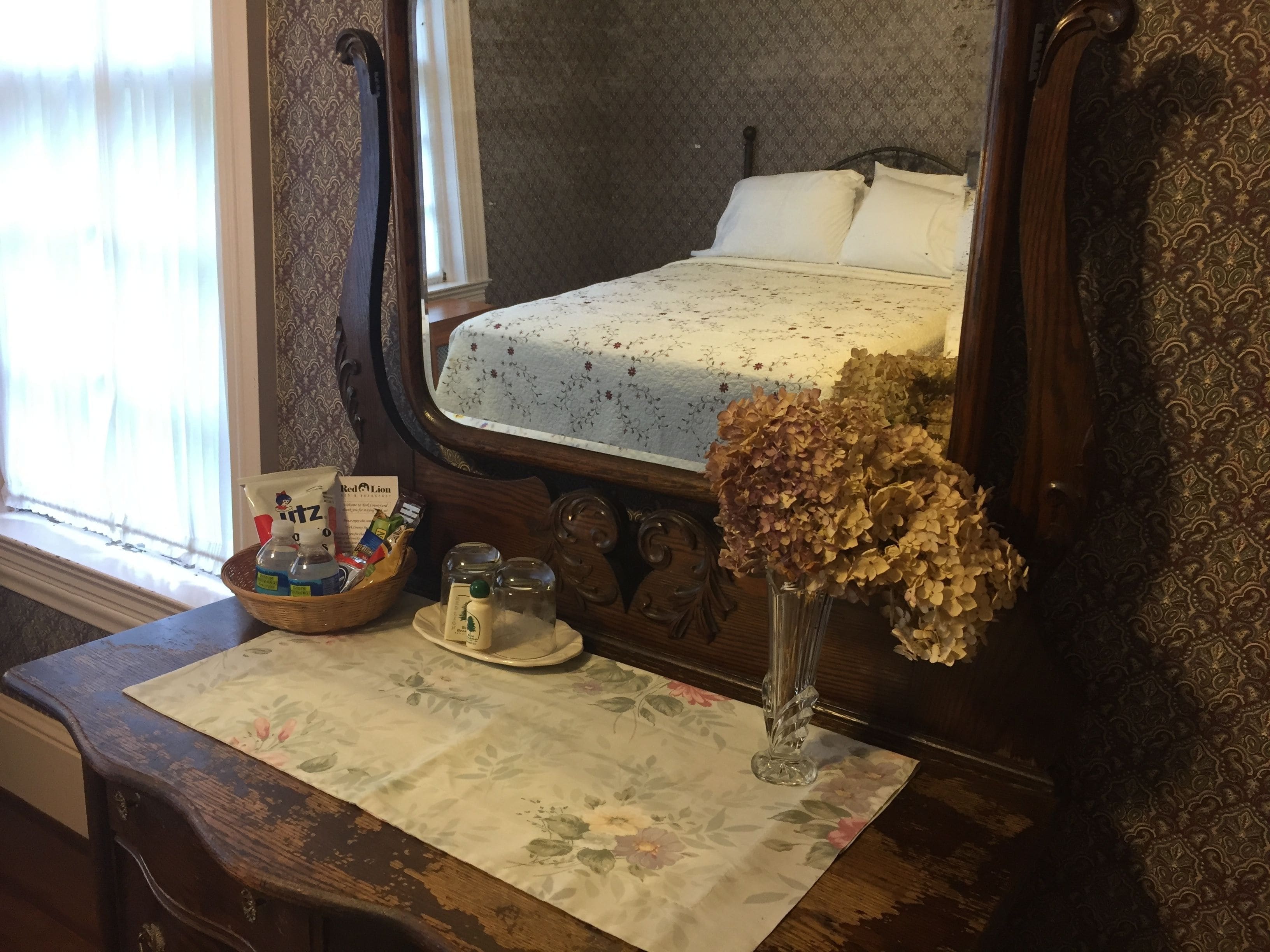 Une chambre à coucher avec un lit, une table de chevet sur laquelle sont posés des articles de toilette, et un vase contenant des fleurs séchées.