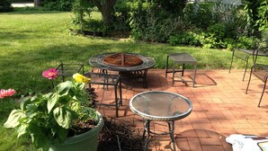 A patio area with a round table, a square table, and a metal chair. There is a potted plant with flowers and a fire pit.