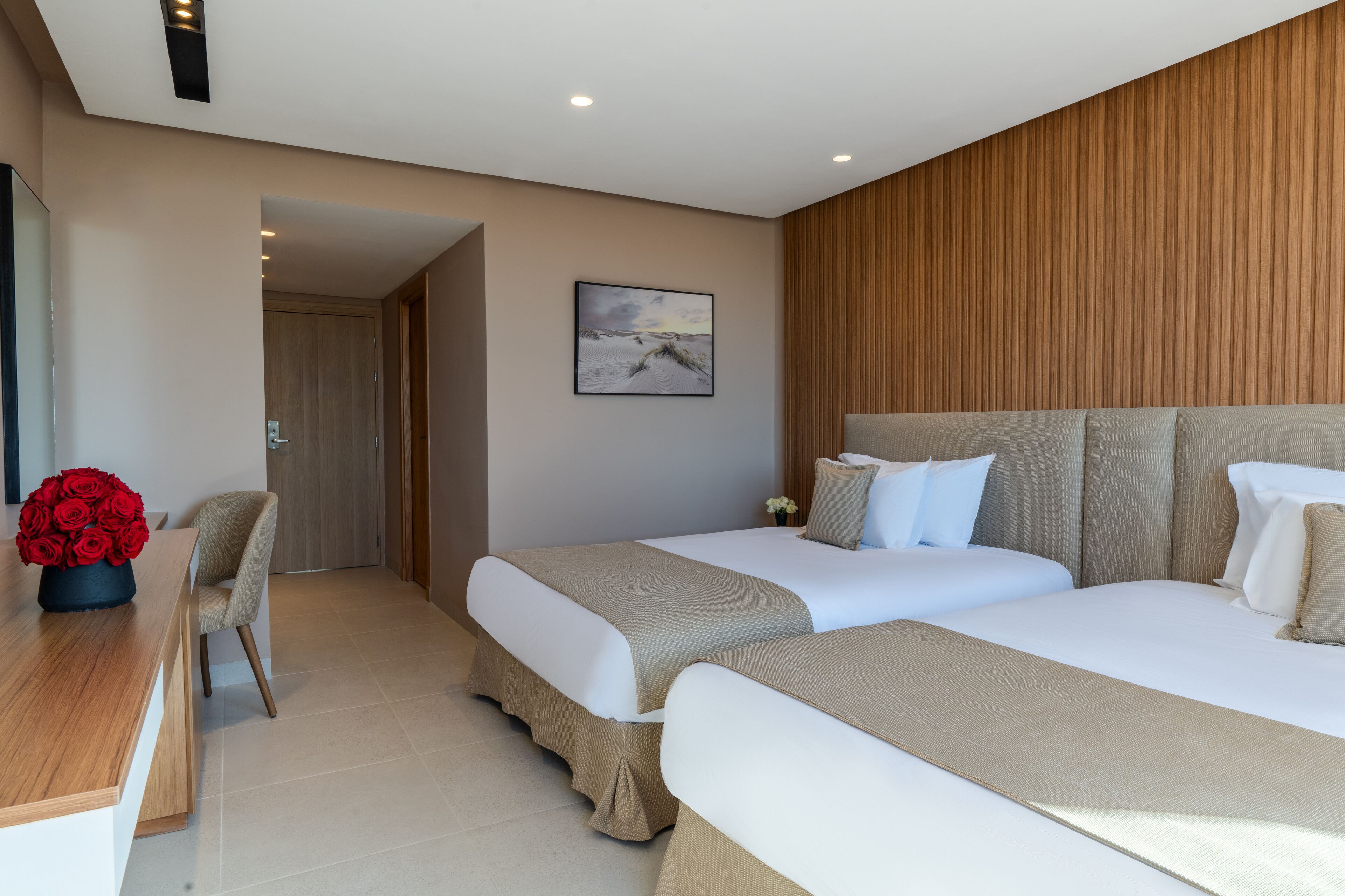 A modern hotel room with two beds, a wooden panel wall, a painting, and a desk with a vase of red roses.