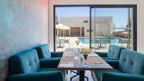 A dining area with teal sofas, a wooden table set for four, and a view of a swimming pool and building through large glass doors. - Kenzi Rissani Hotel (Errachidia)