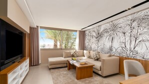 A modern living room with a large flat-screen TV, a beige sofa, a coffee table, and a mural of tropical plants on the wall. - Kenzi Rissani Hotel (Errachidia)