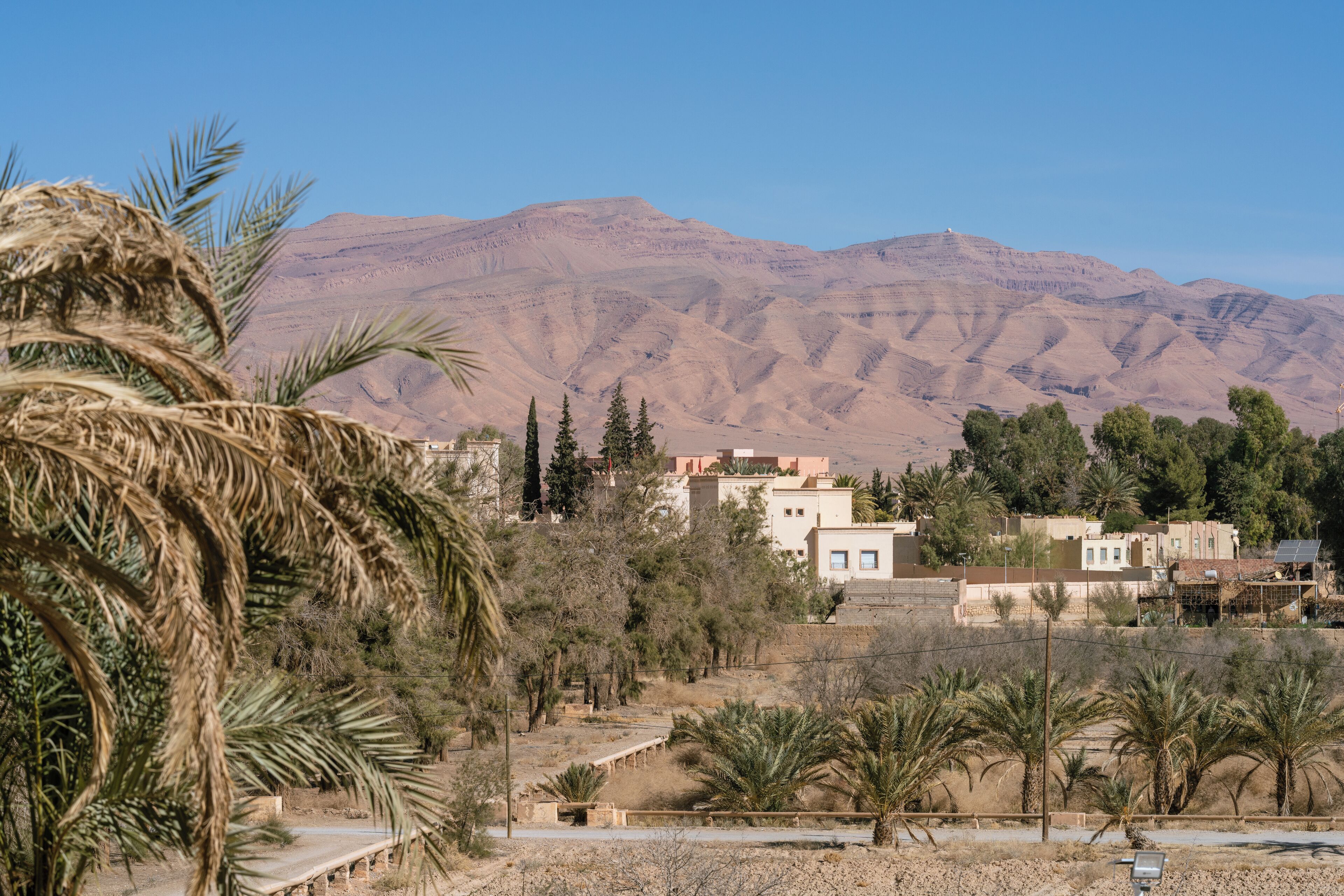 Ένα έρημο τοπίο με βουνά στο βάθος, φοίνικες και κτίρια, με καθαρό ουρανό.