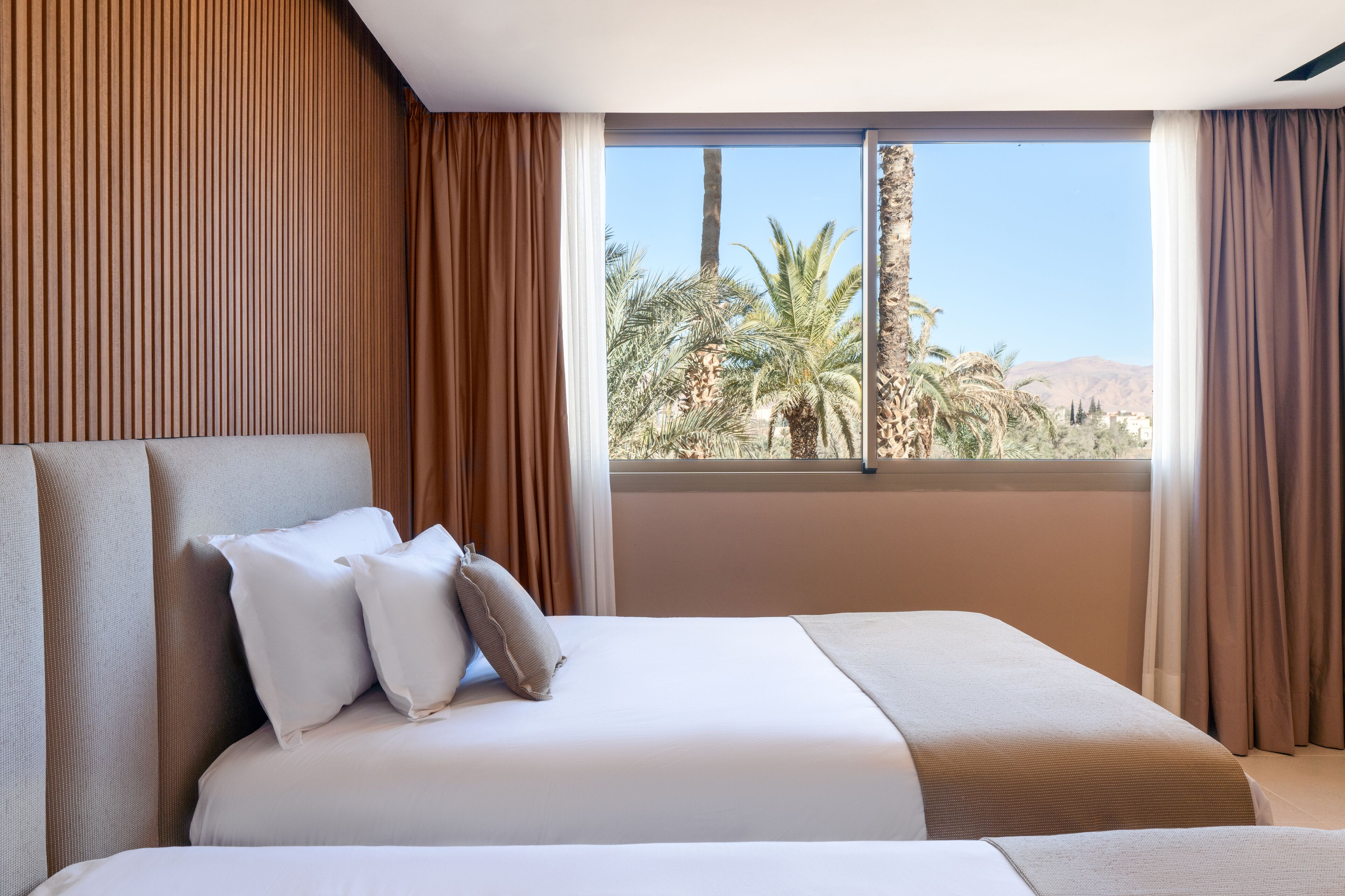 A hotel room with a large bed, a view of palm trees and mountains, and brown curtains.