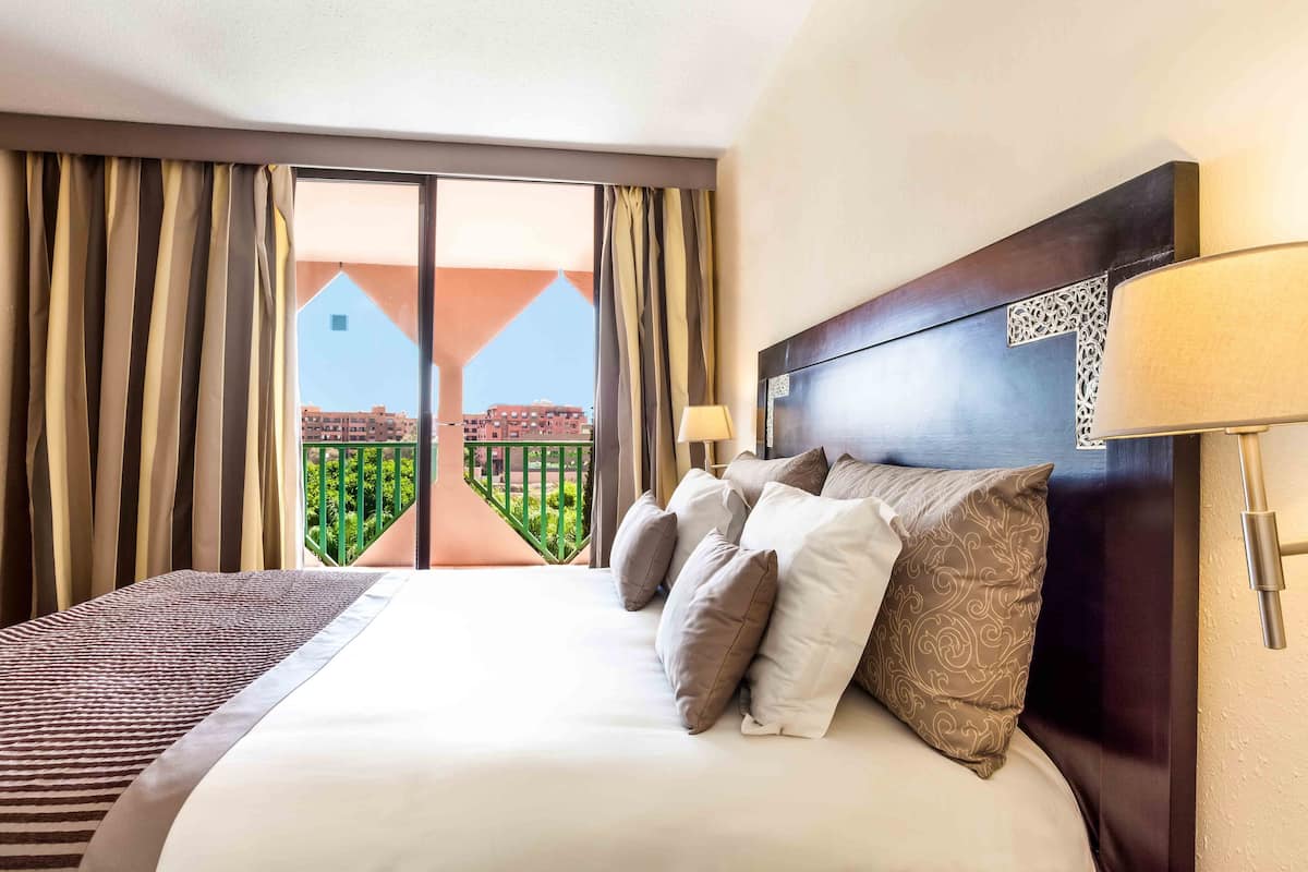 Habitación de hotel con una cama grande, varios almohadones, cabecera con panel decorativo, ventana con cortinas y vistas a edificios y áreas verdes.