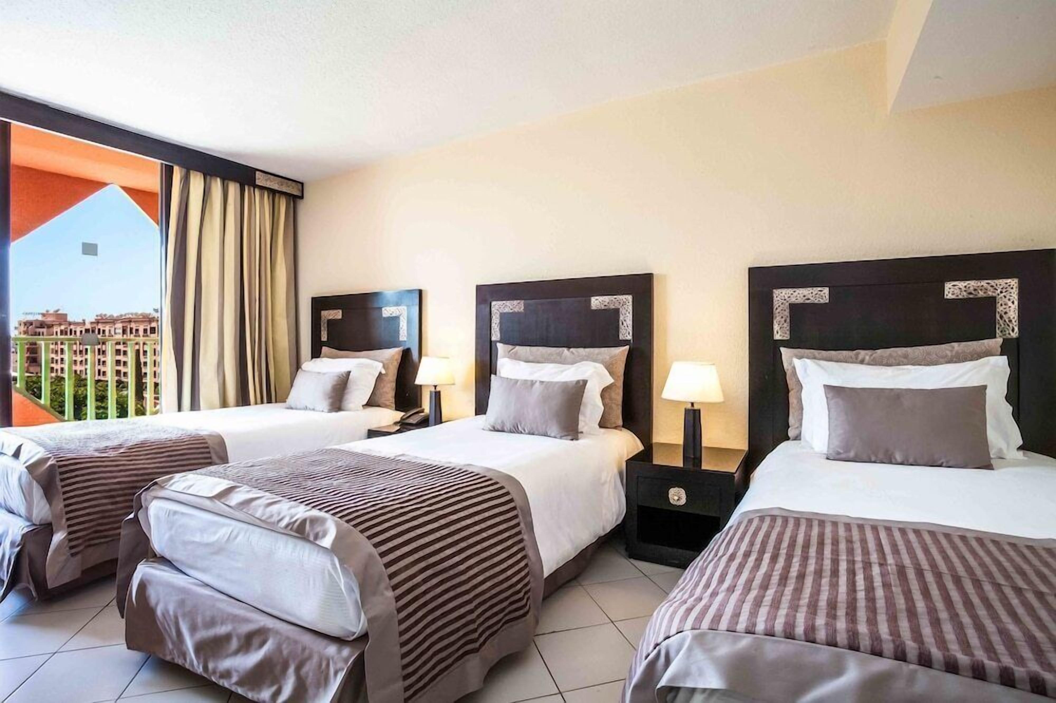 A hotel room with three beds, a balcony with curtains, and a view of buildings.