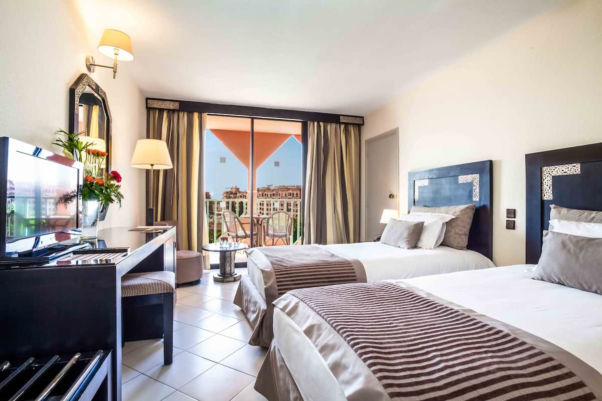 Habitación de hotel con dos camas, un escritorio con ordenador, un piano y un balcón con vistas a edificios.