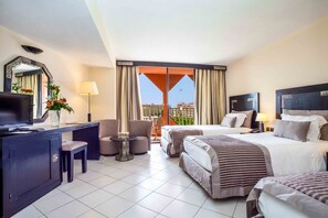 A hotel room with a large bed, a desk with a TV, a chair, a small table with a lamp, and a view of the city through a large window. - Atlas Asni (Marrakech)