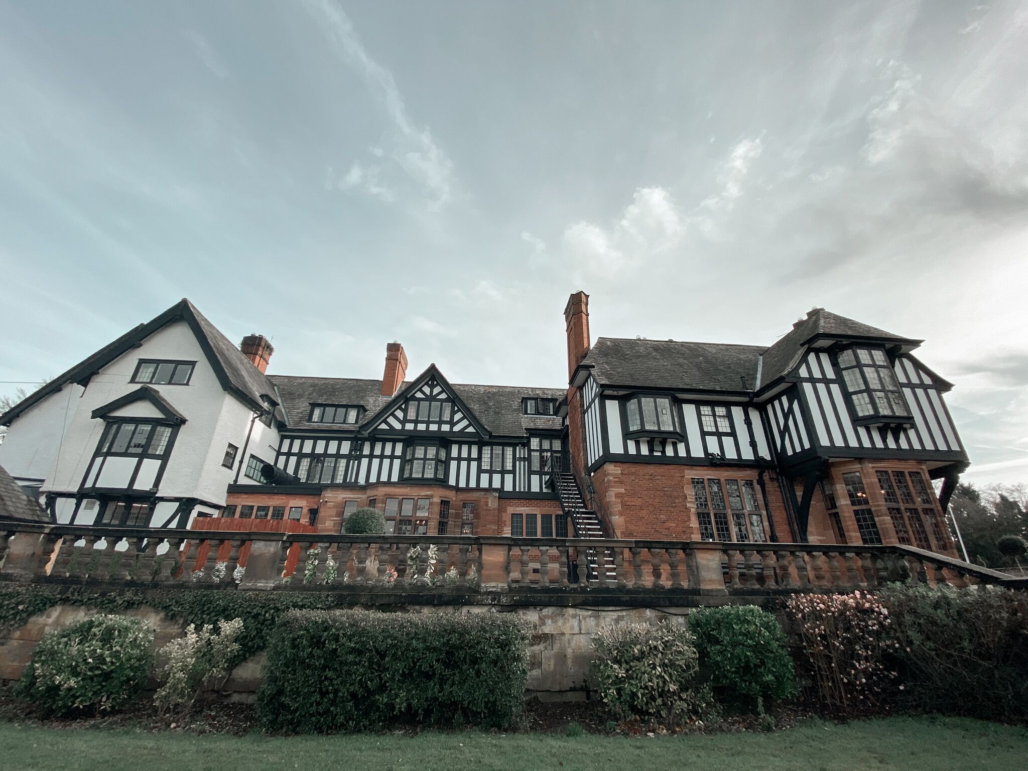 A traditional Tudor-style building with multiple chimneys, a stone wall with a balustrade, and a well-maintained garden.