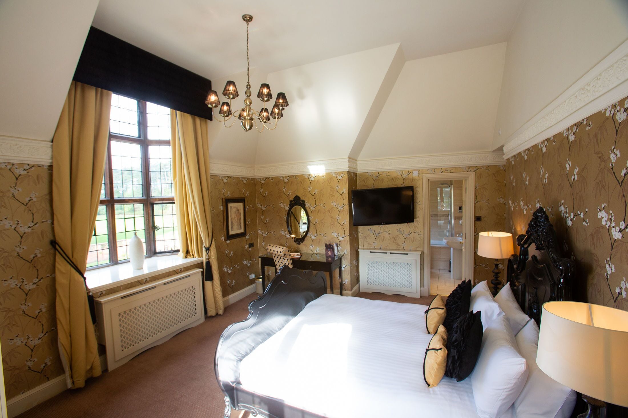 A hotel room with a large bed, a chandelier, a television, and a window with curtains.