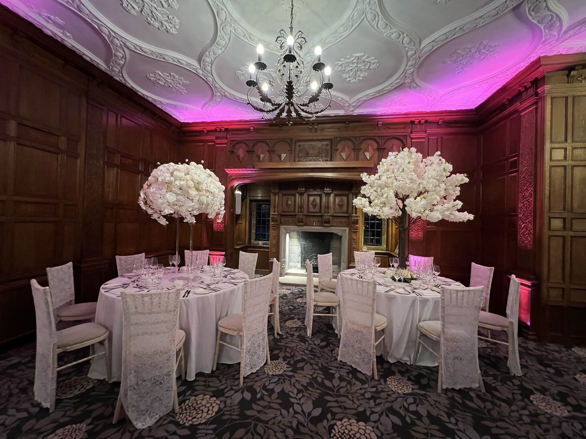A formal dining room with round tables, white lace chair covers, and large white floral centerpieces.