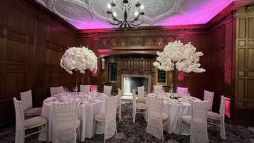 A formal dining room with round tables, white lace chair covers, and large white floral centerpieces.