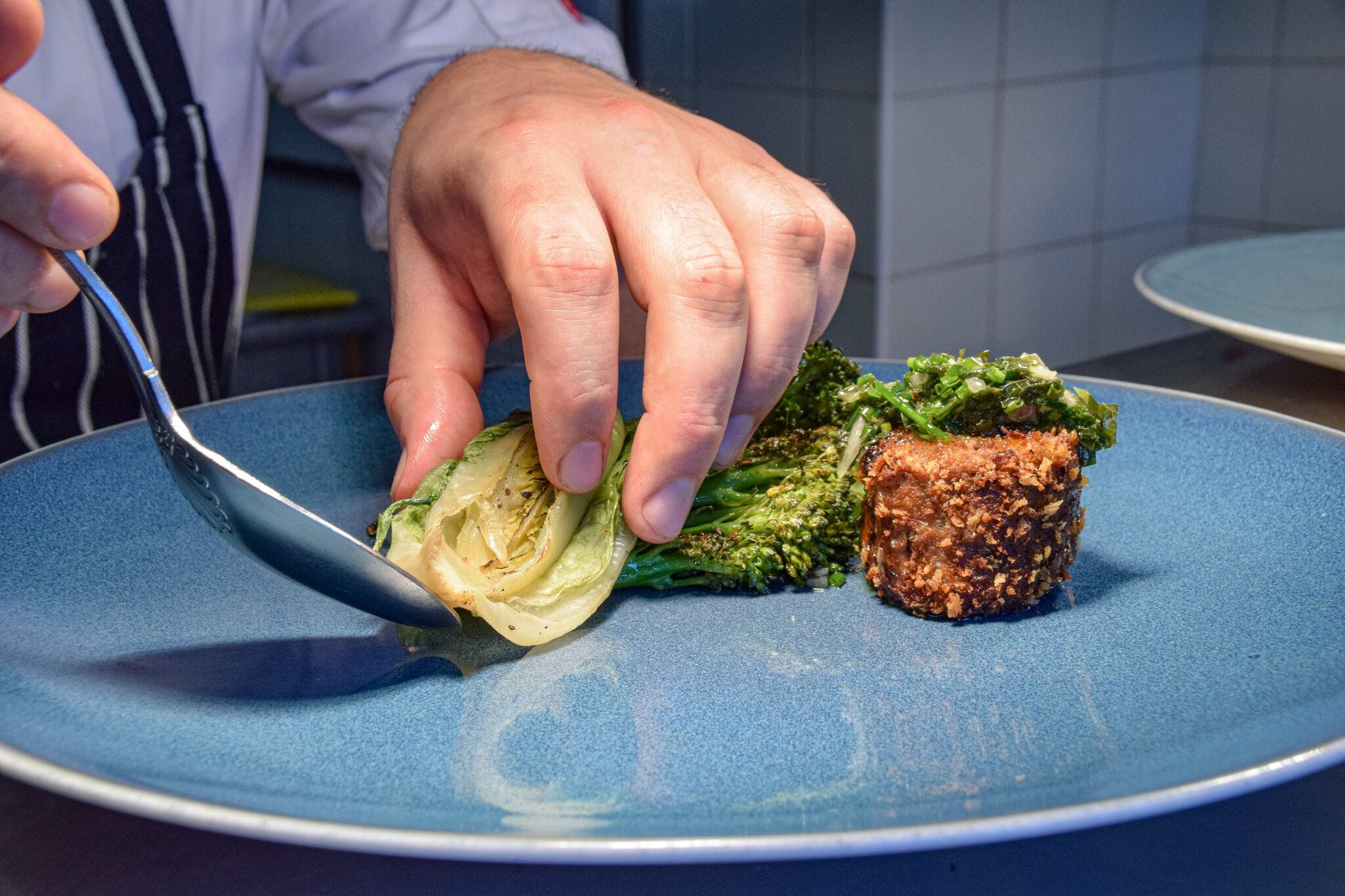 Una mano che tiene un cucchiaio sopra un piatto blu con un fiore di broccolo e un impanatura.