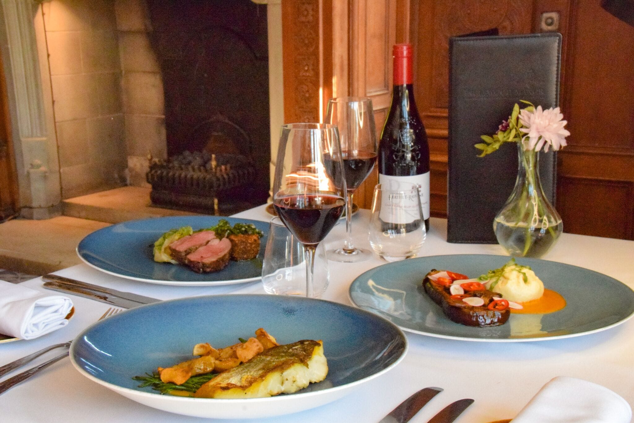 A dining table with three plates of food, a bottle of wine, and a vase with flowers.