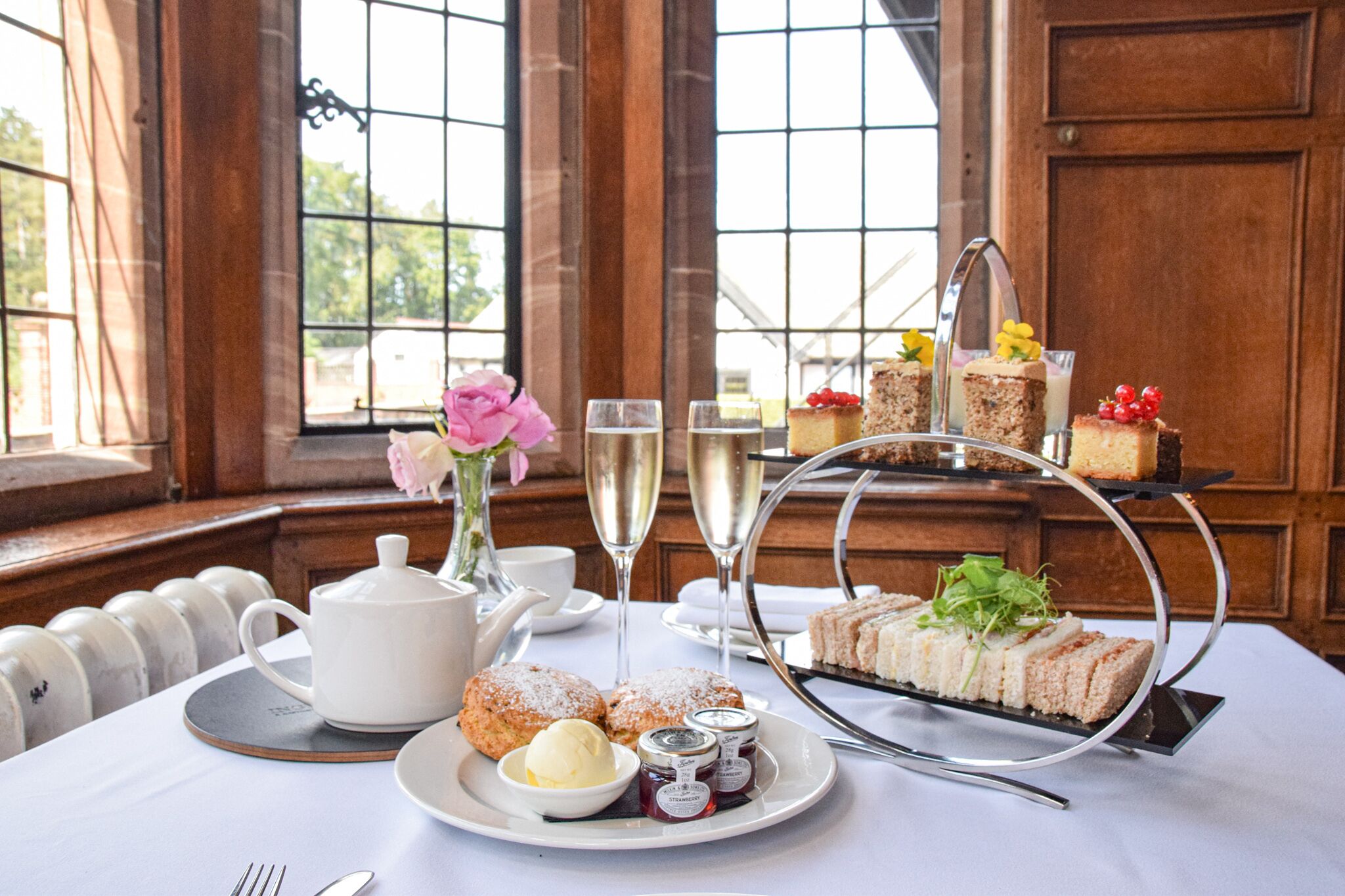 A table set with a teapot, cups, a tiered dessert stand, pastries, and a vase of flowers.