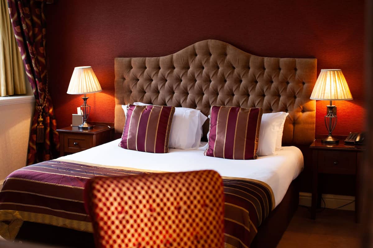 A hotel room with a large bed, two bedside tables with lamps, a patterned armchair, and a window with curtains.