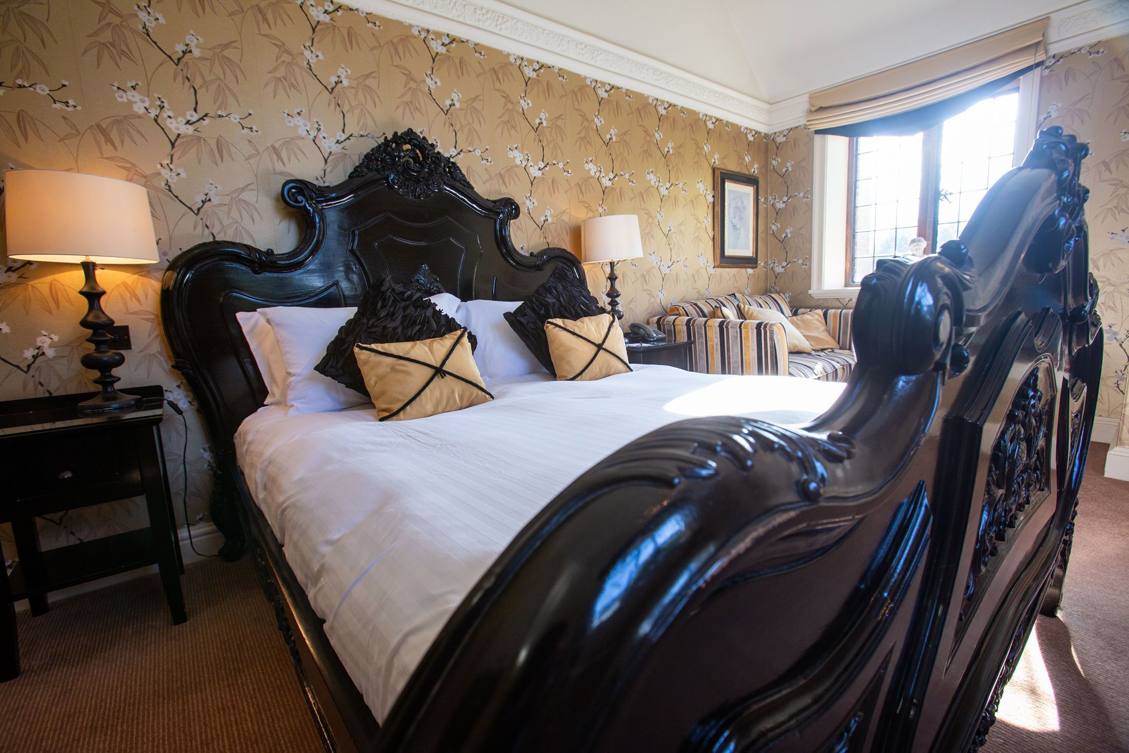 A classic four-poster bed with a dark finish, a white bedspread, and decorative pillows. There is a nightstand with a lamp to the left of the bed, and a window with curtains to the right.