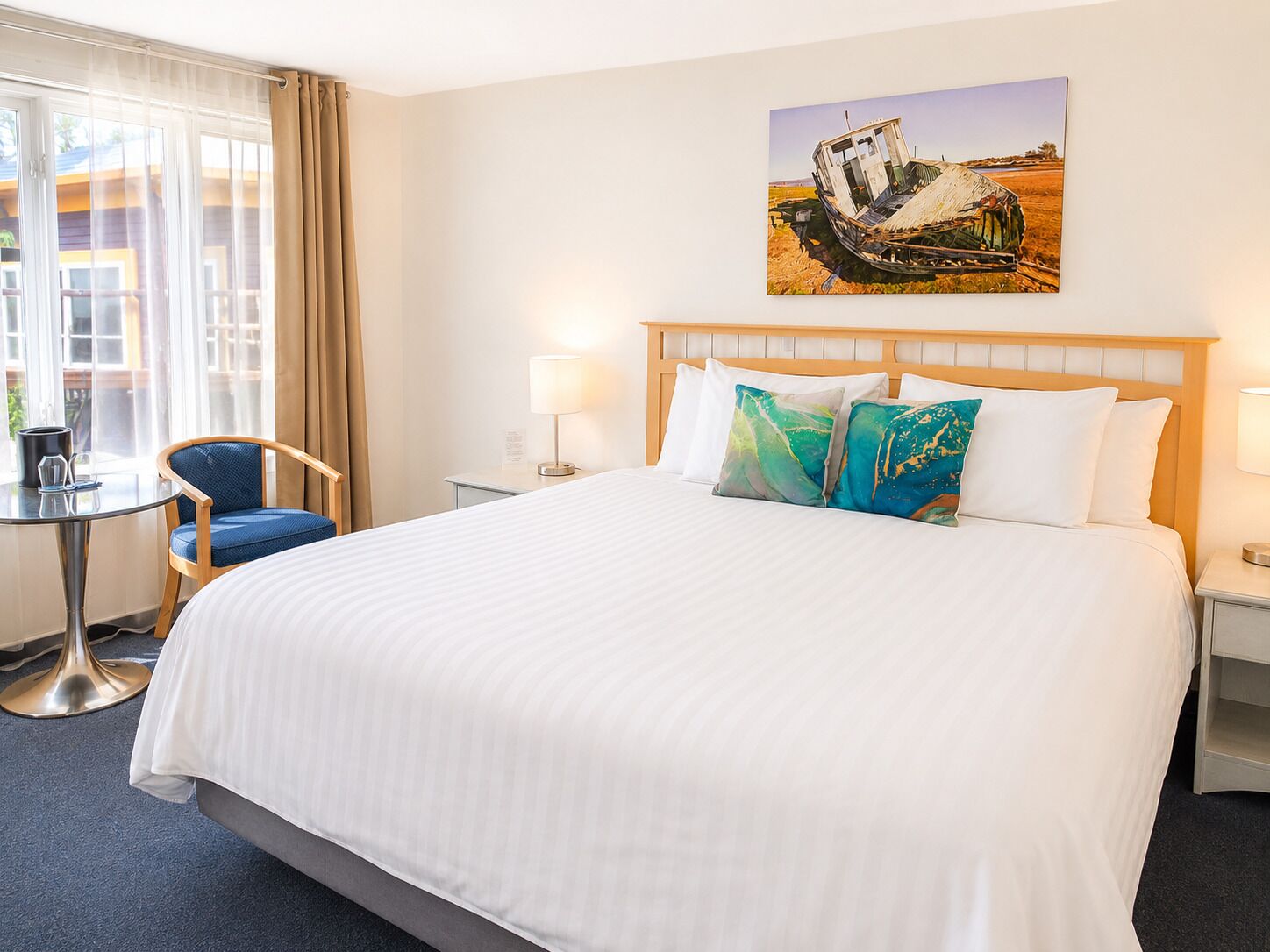 A hotel room with a bed, a chair, a small table, and a painting on the wall.
