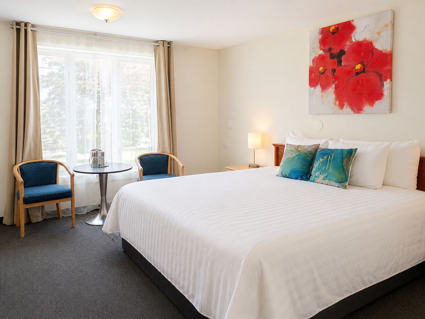 A hotel room with a bed, two chairs, a small table, a lamp, and a painting on the wall.