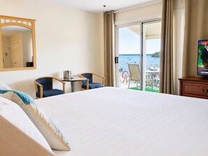 A hotel room with a bed, two chairs, a small table, a television, and a view of the sea.