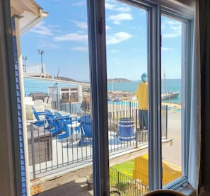 Une vue à travers une fenêtre sur un espace piscine, avec des chaises bleues, un parasol jaune et une jetée où sont amarrés des bateaux.