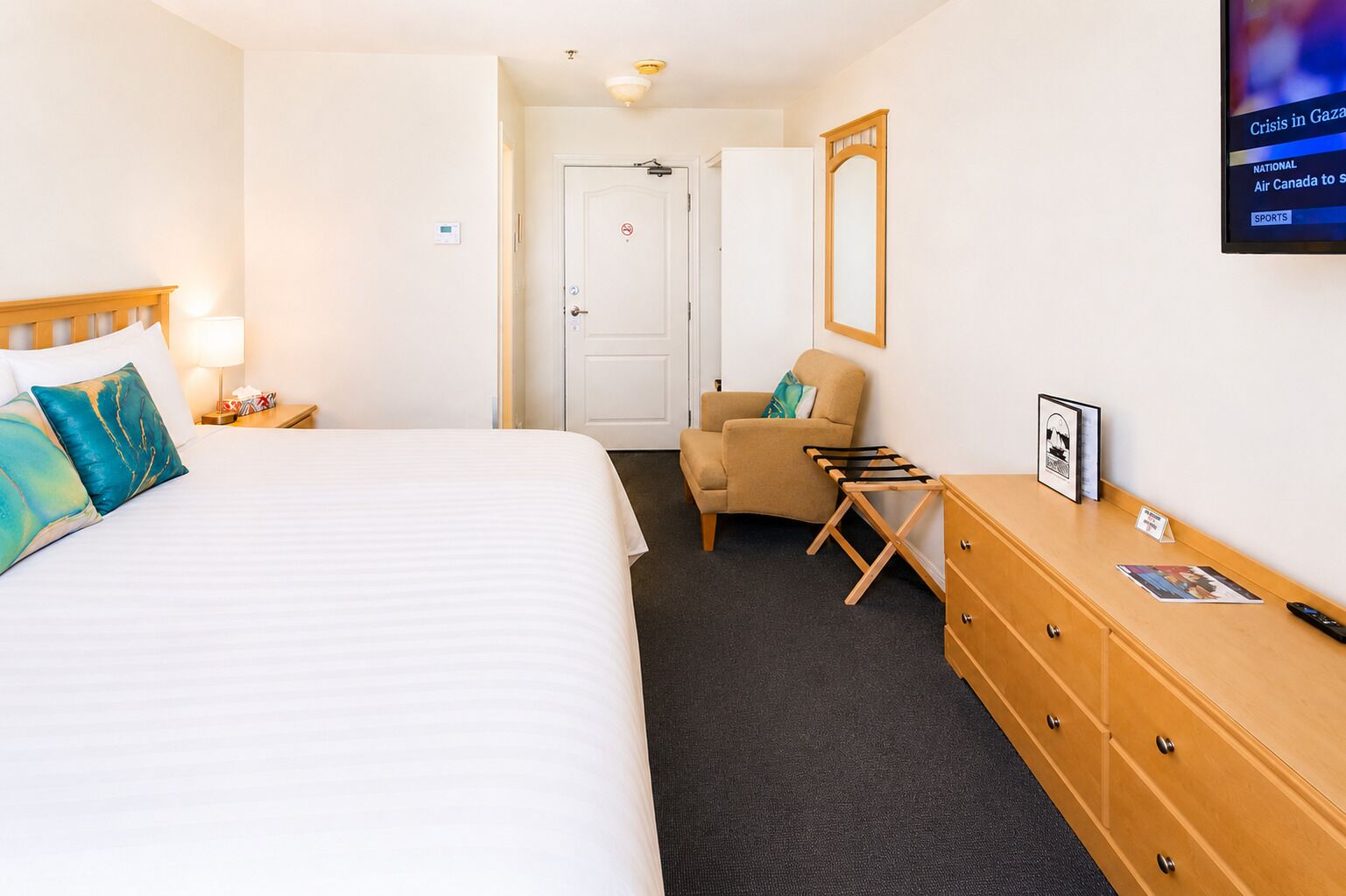 Ein Hotelzimmer mit einem großen Bett, einem Sessel, einem Schminktisch und einem Fernseher, der Nachrichten zeigt.