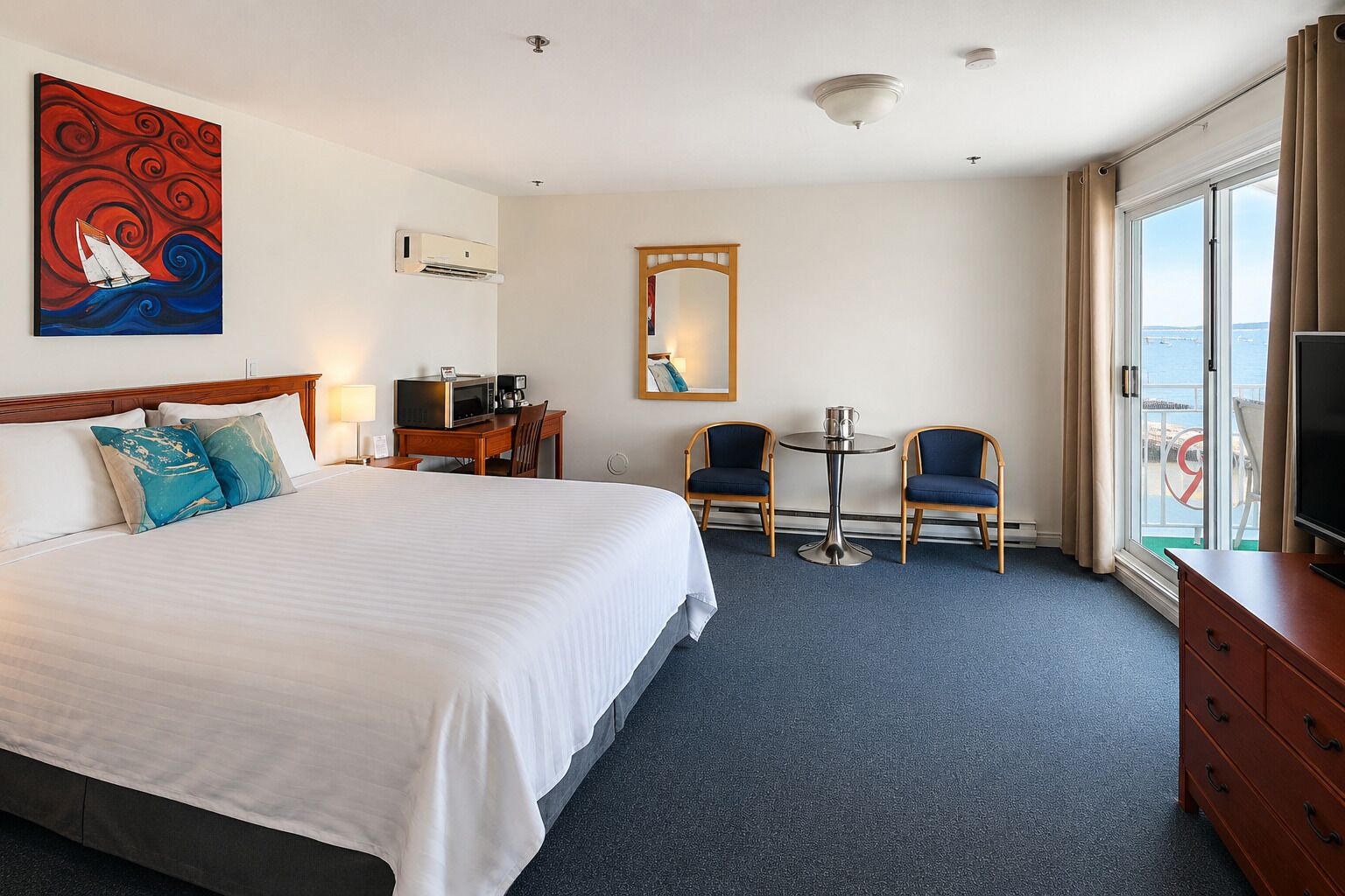 A hotel room with a bed, desk, chairs, a TV, and a view of the sea.