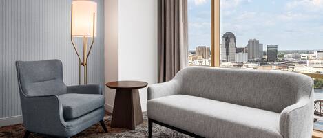 Habitación de hotel moderna con un sillón gris, un sofá, una lámpara de pie y vistas a la silueta urbana a través de amplias ventanas.