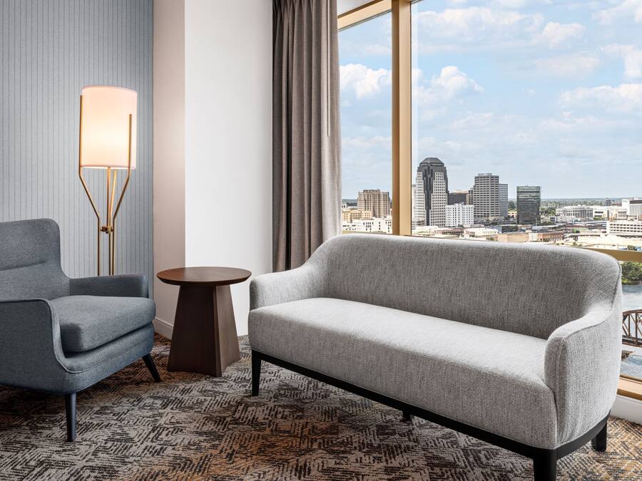 A modern hotel room with a grey armchair, a sofa, a floor lamp, and a view of the city skyline through large windows.
