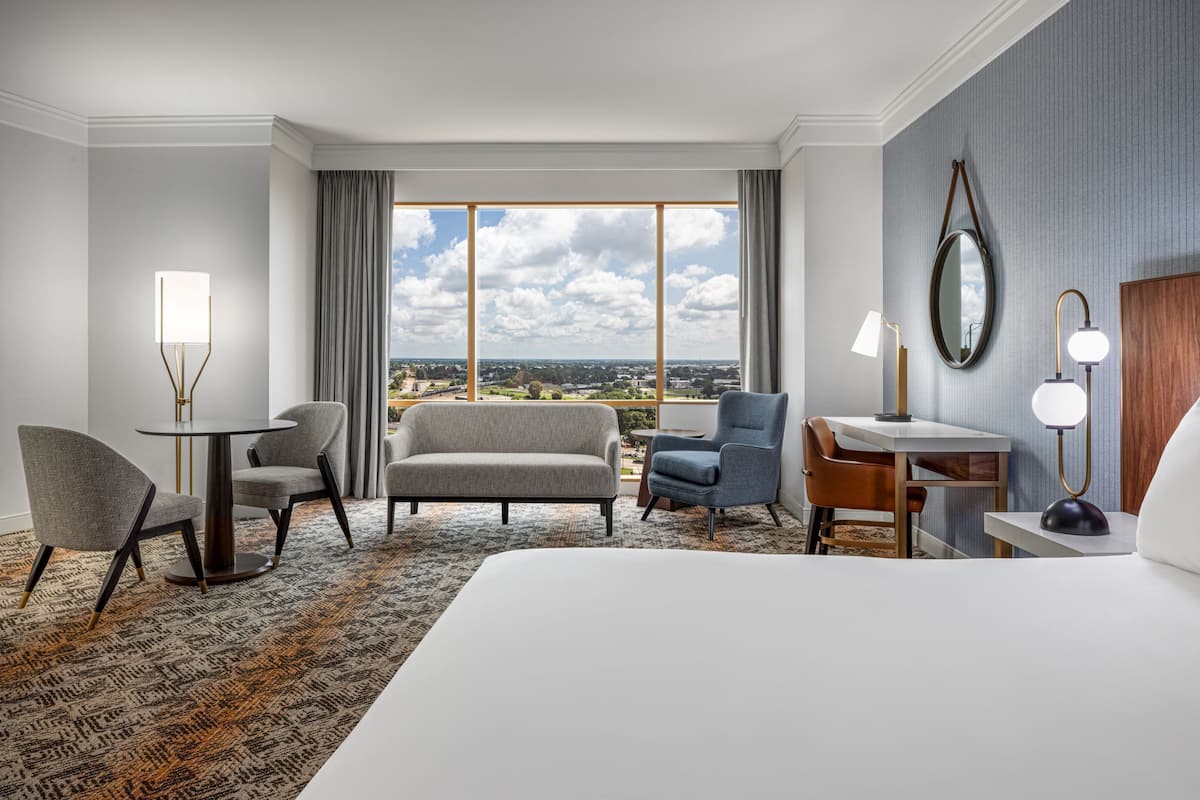 Habitación de hotel con una cama grande, un escritorio, dos sillones, una mesita y vistas a la ciudad a través de amplios ventanales.