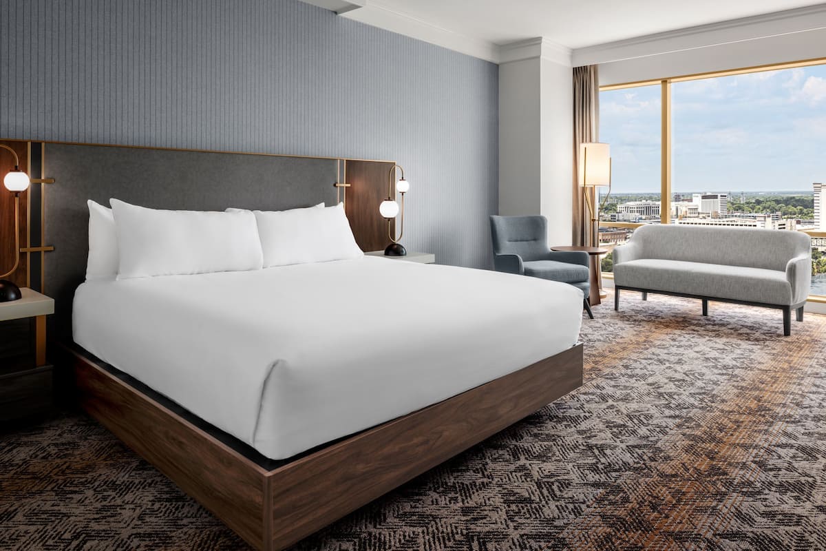 Habitación de hotel con una cama grande, un escritorio con lámpara, una silla y vistas a la ciudad.