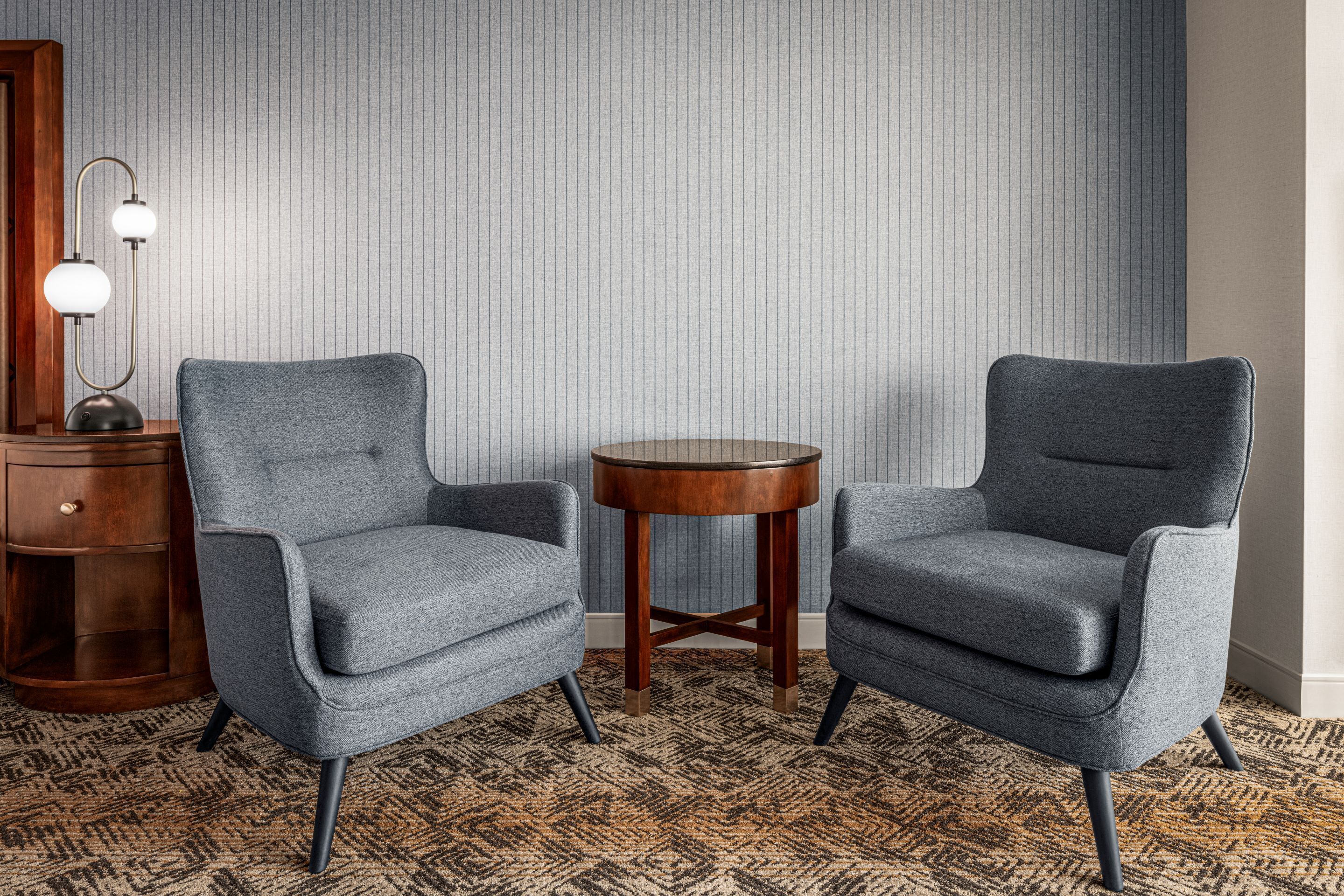 Deux fauteuils gris aux pieds en bois et une petite table en bois placée entre eux, le tout posé sur un tapis à motifs.