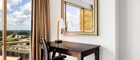 A hotel room with a large window, a desk, a chair, and a mirror.