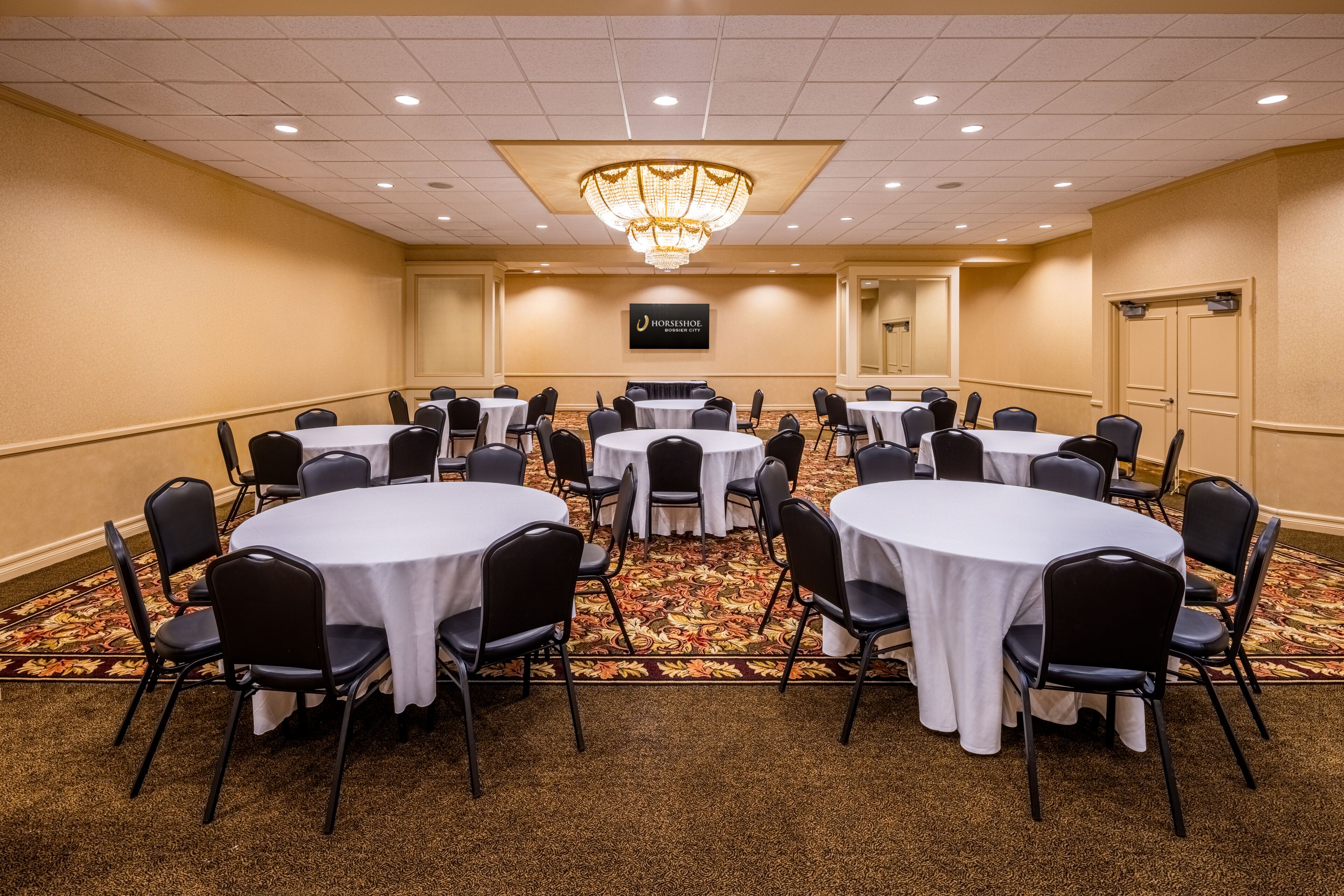 A conference room with multiple round tables, black chairs, and a chandelier.