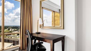 A hotel room with a large window, a desk, a chair, and a mirror.