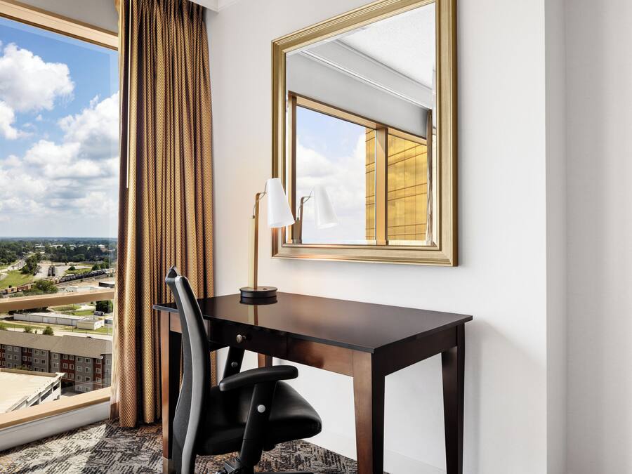A hotel room with a large window, a desk, a chair, and a mirror.