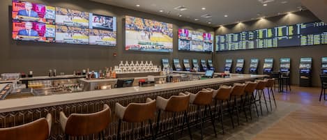 A bar area with multiple screens displaying sports events, a bar counter with various bottles, and several bar stools.