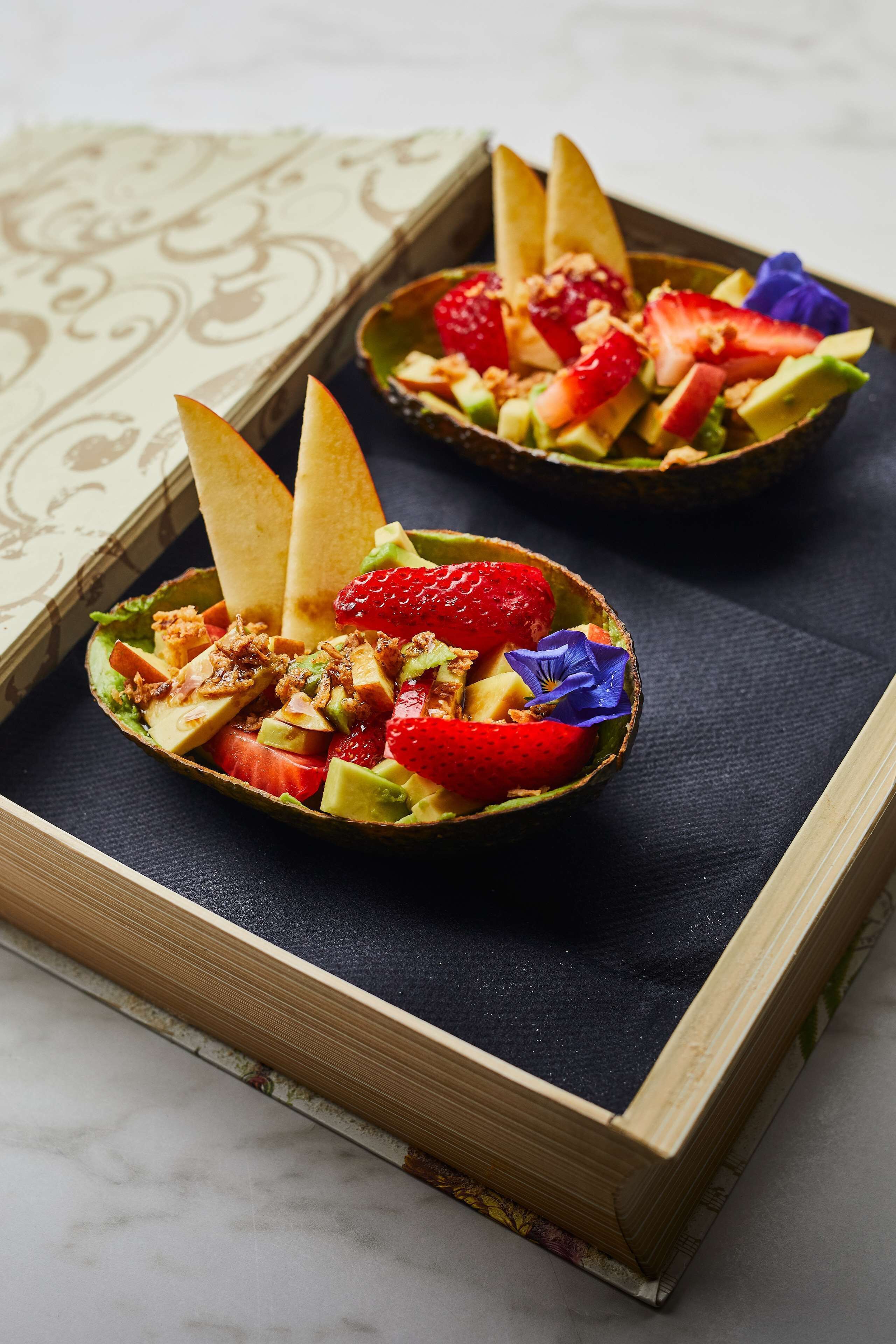 Two bowls of fruit salad with a decorative flower, placed on a dark cloth inside a wooden box.