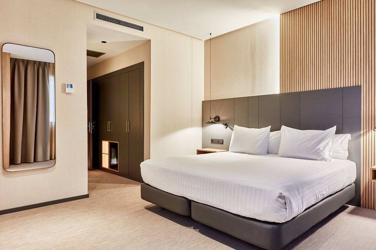 Ein modernes Hotelzimmer mit einem großen Bett, einem Nachttisch mit Lampe, einem Kleiderschrank und einem Spiegel.