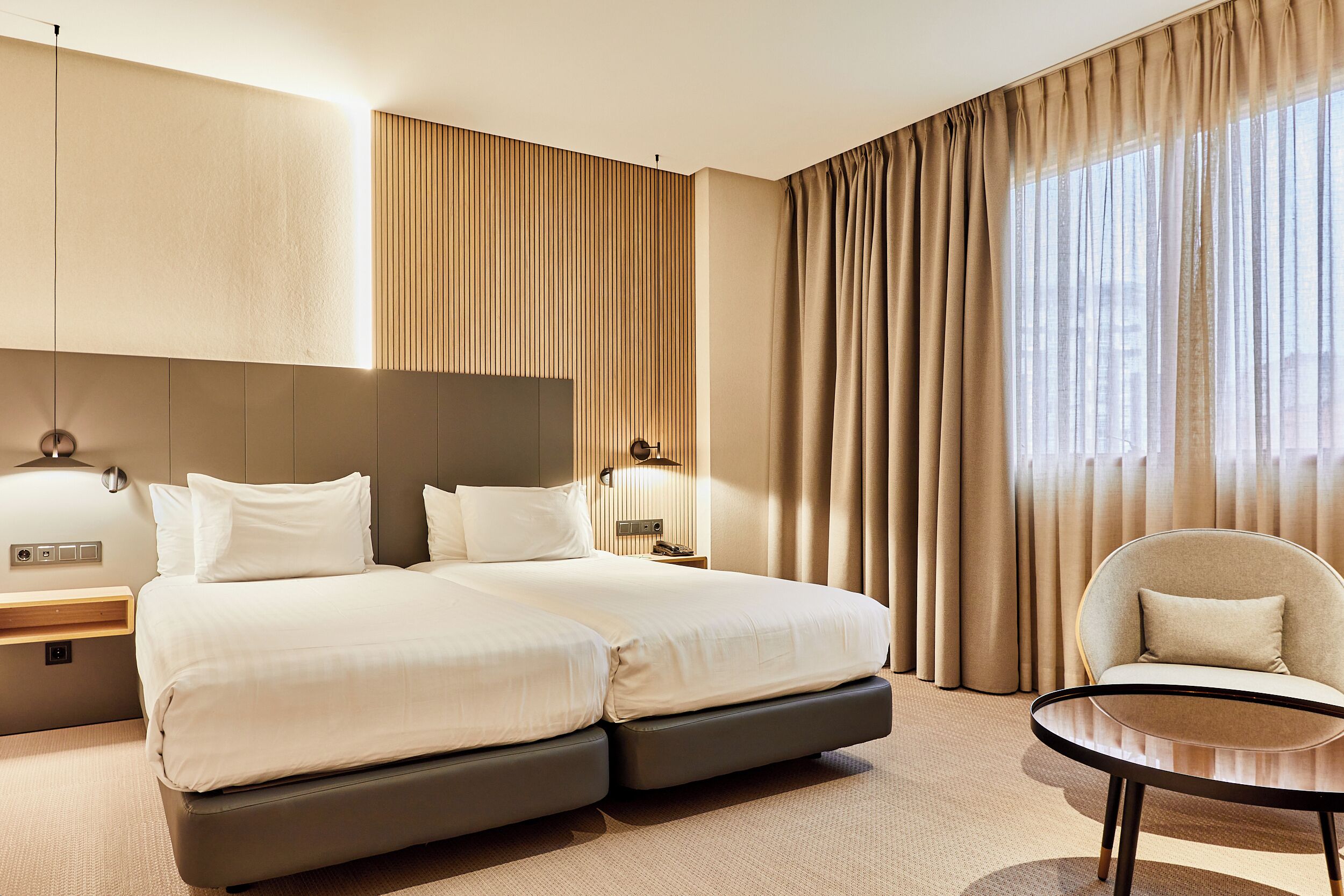 Une chambre d’hôtel moderne avec un grand lit, une table de chevet, une chaise et une vue sur la ville grâce à de grandes fenêtres.