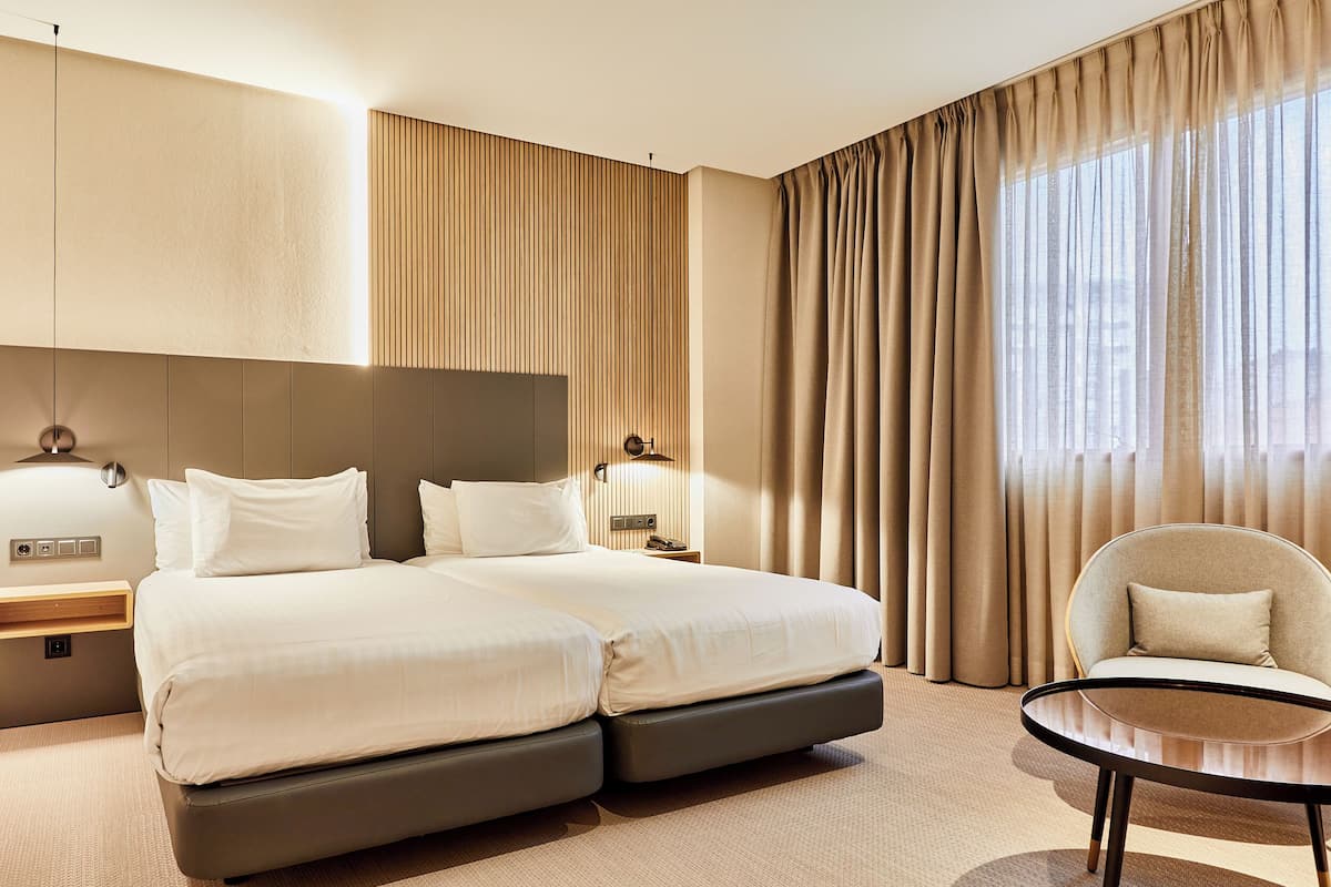 A modern hotel room with a large bed, a bedside table, a chair, and a view of the city through large windows.