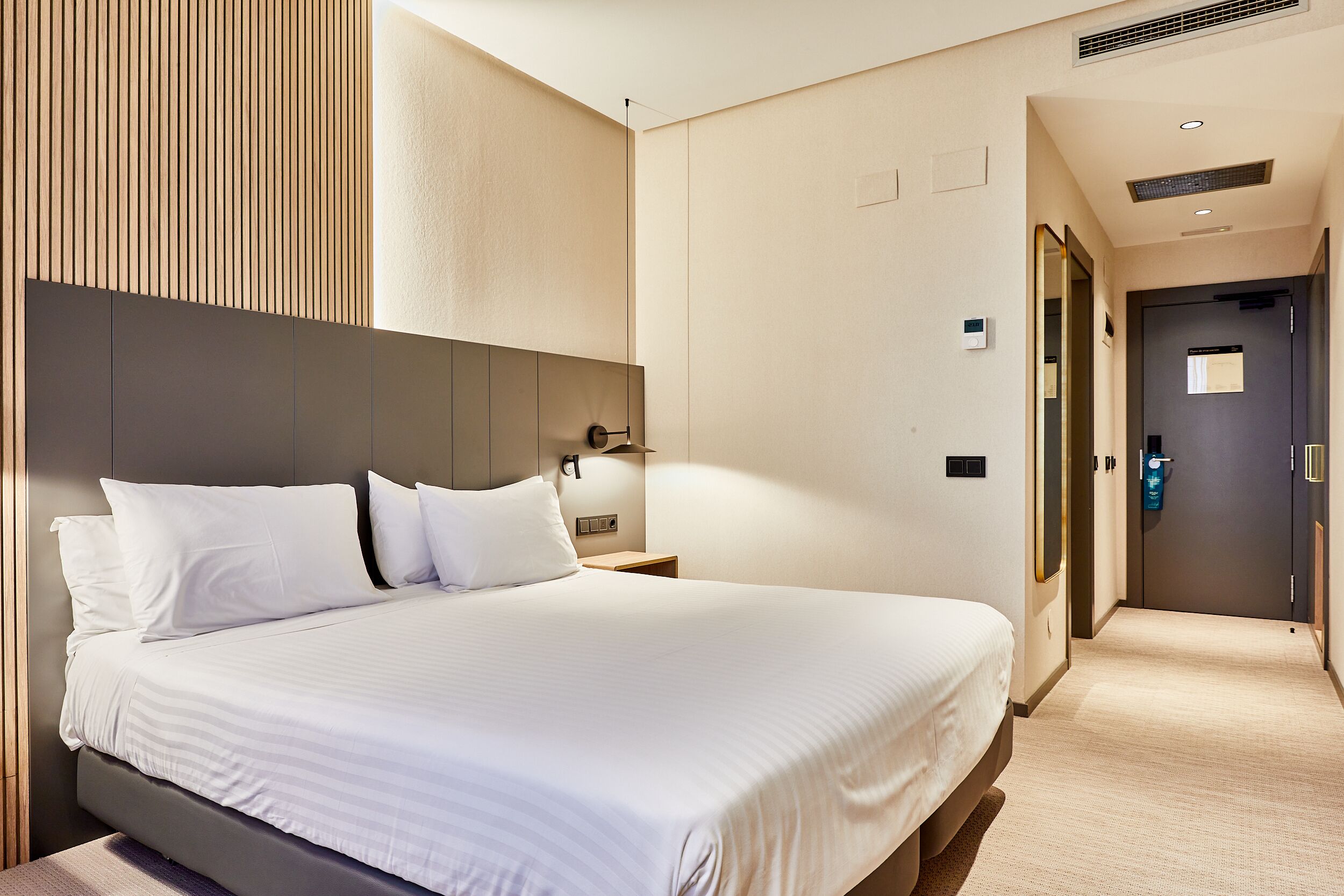 Habitación de hotel con una cama grande, una mesita de noche, una lámpara de pared y una puerta que da a otra habitación.