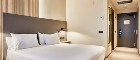 Habitación de hotel con una cama grande, una mesita de noche, una lámpara de pared y una puerta que da a otra habitación.