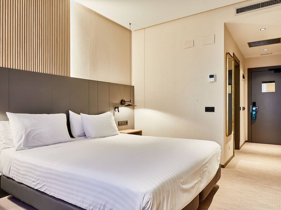 Habitación de hotel con una cama grande, una mesita de noche, una lámpara de pared y una puerta que da a otra habitación.