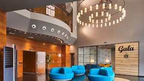 A modern hotel lobby with a curved ceiling, a central chandelier, and a reception area with a 'Gala' sign.