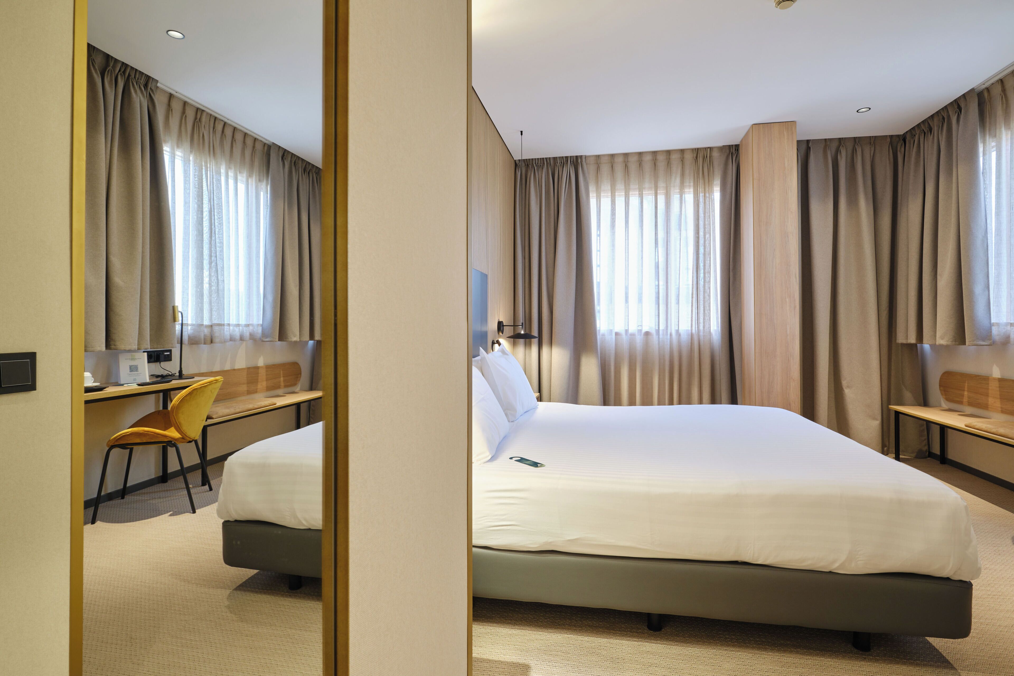 A hotel room with a bed, a desk, a chair, and a large window with curtains.
