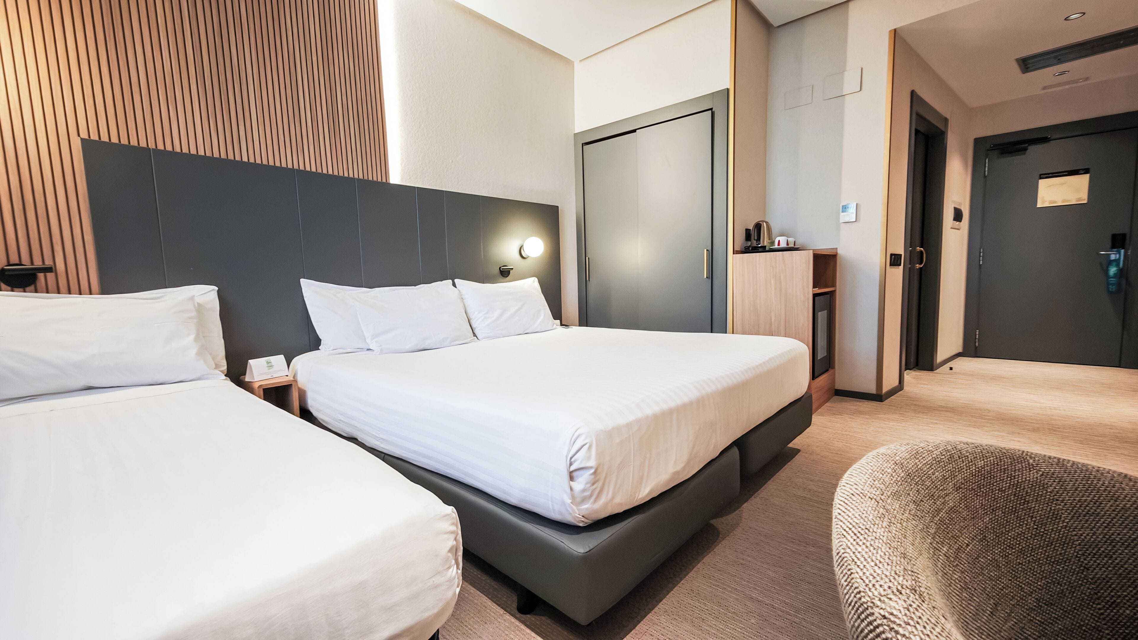 A hotel room with two beds, a dark headboard, a wooden accent wall, a wardrobe, and a small seating area.