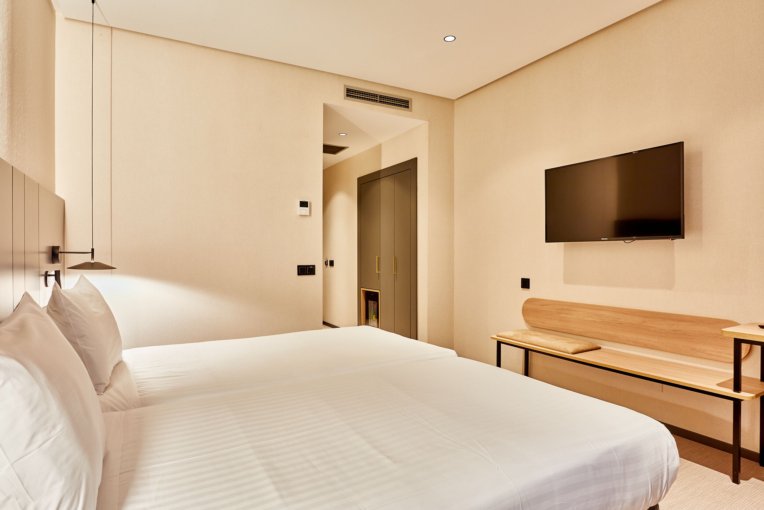 A modern hotel room with a large bed, a flat-screen TV mounted on the wall, and a small wooden bench.